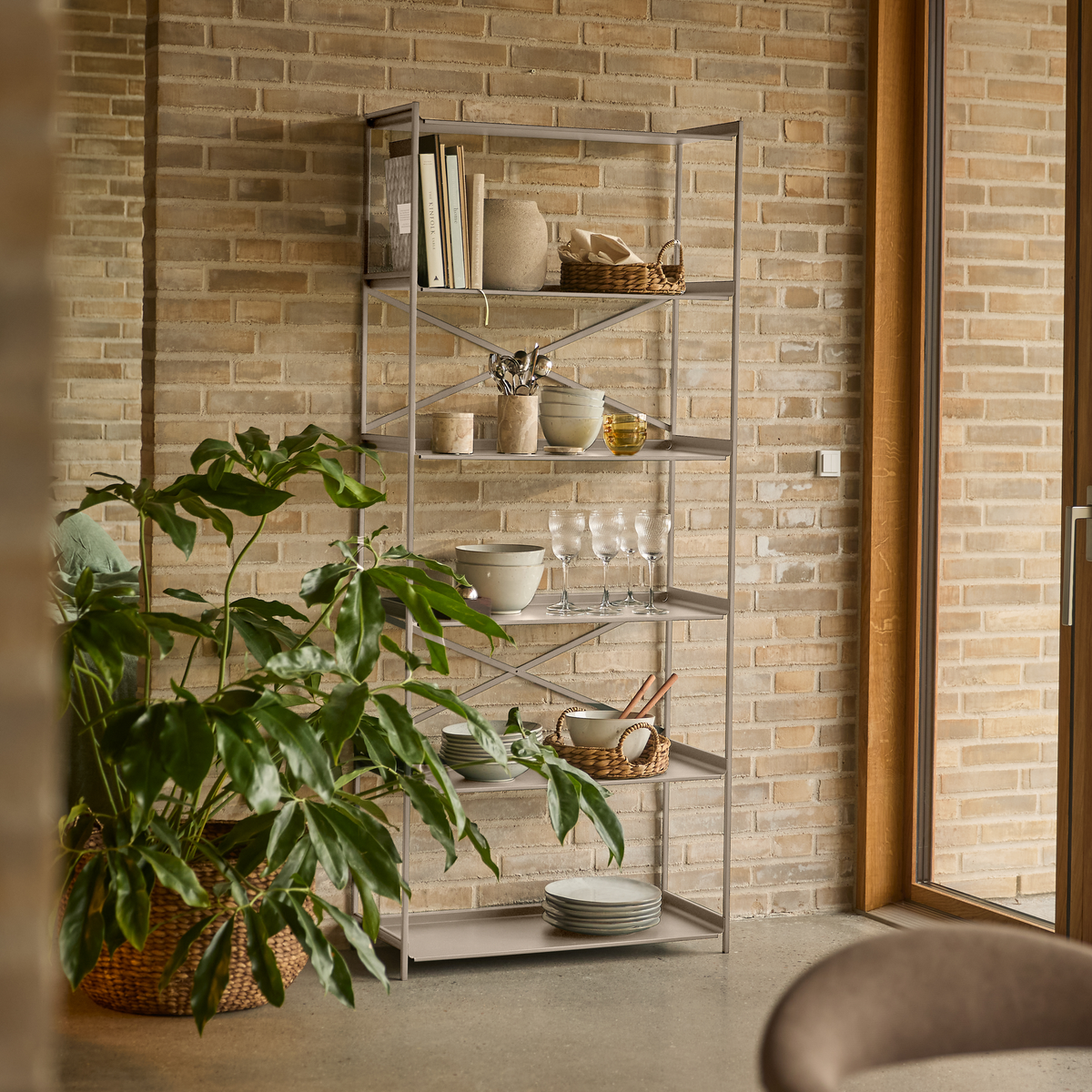 Åben metalreol mod lys murstensvæg med keramik, glas, bøger og kurve; stor grøn potteplante ved siden af et vindue.