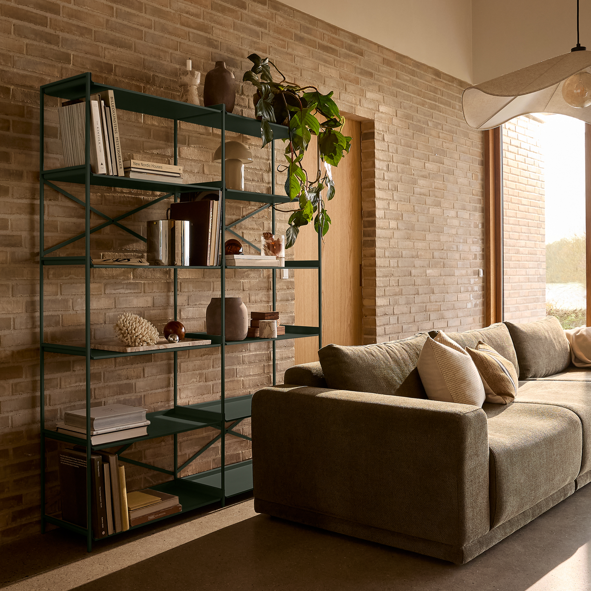 Stue med grøn metalreol med bøger (bl.a. New Nordic Houses), keramik og hængeplante, beige murstensvæg og stor grå sofa med puder i blødt dagslys fra stort vindue.