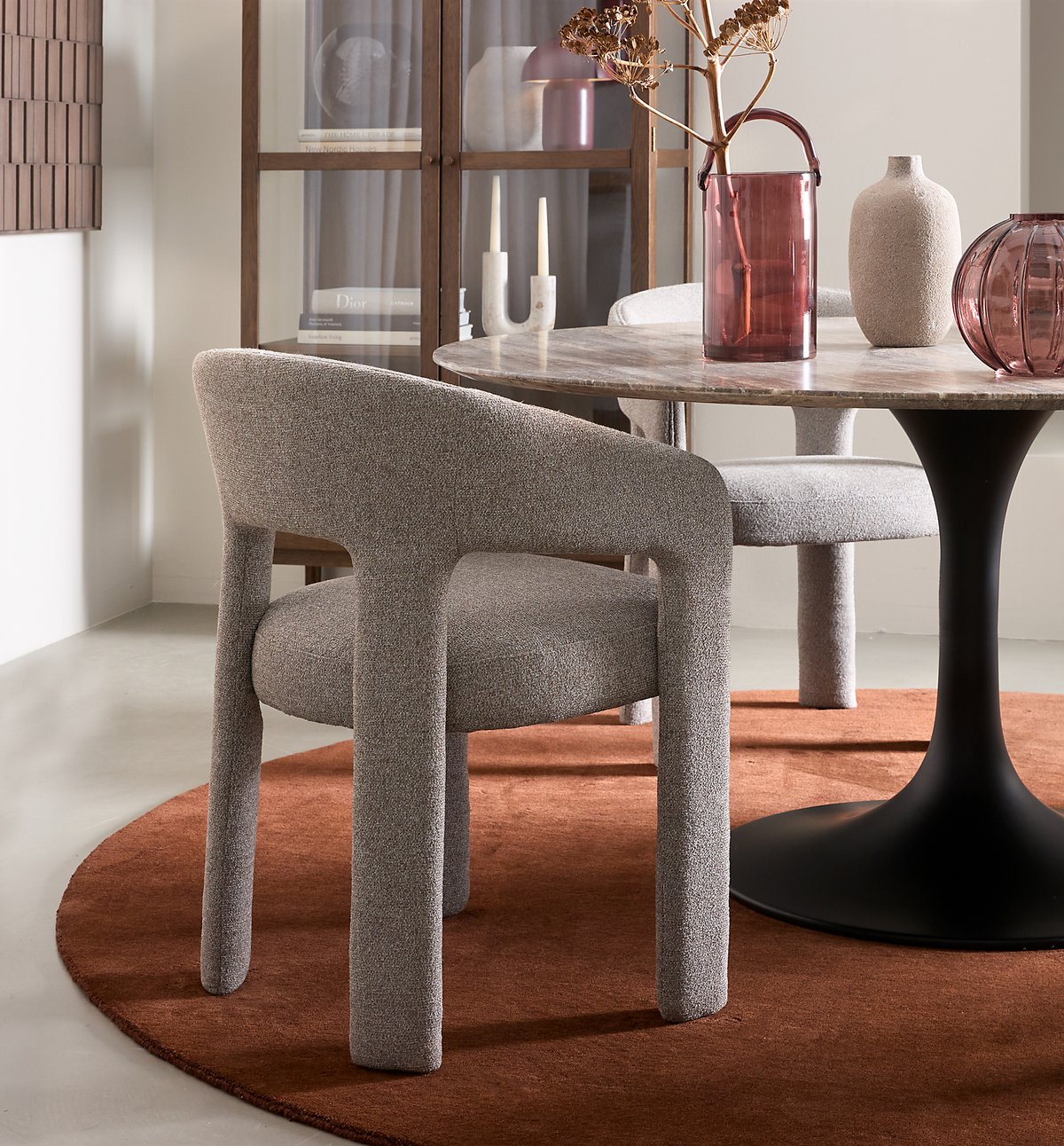Moderne spiseplads med grå boucléstol, rundt bord med stenplade og sort trompetfod på et rustfarvet rundt tæppe, pyntet med rosa glasvaser og lysestager.