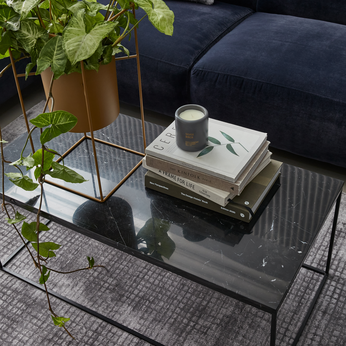 Sort marmorbord med grøn potteplante i guldfarvet stativ, stak designbøger (Cereal, Kinfolk, A Frame for Life, The Design House Book, New Perspectives) og duftlys ”Silver Birch” på sofabord.
