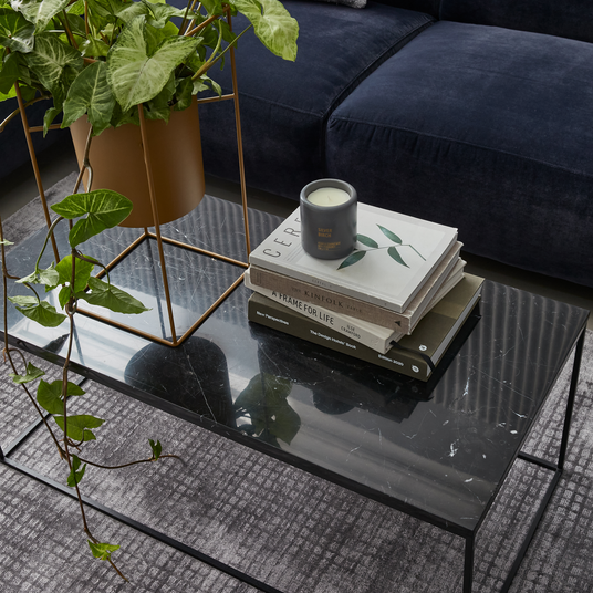 Sort marmorbord med grøn potteplante i guldfarvet stativ, stak designbøger (Cereal, Kinfolk, A Frame for Life, The Design House Book, New Perspectives) og duftlys ”Silver Birch” på sofabord.