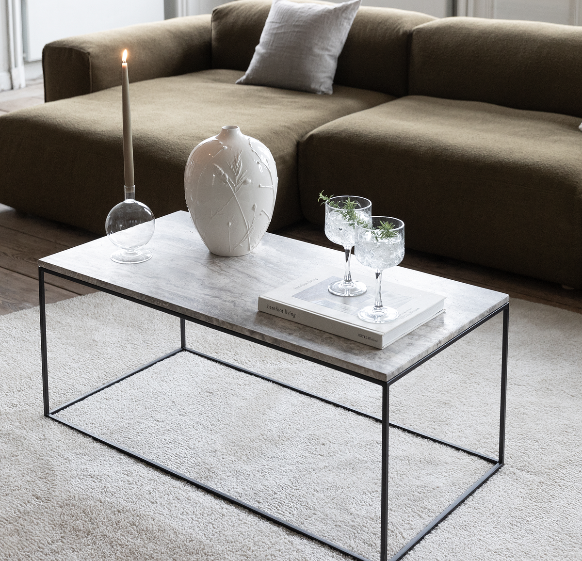 Marmorbord med hvid vase, tændt lys og to glas oven på bogen ”barefoot living” foran en olivengrøn sofa.