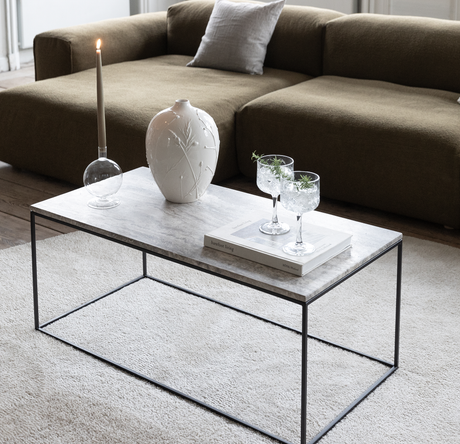 Marmorbord med hvid vase, tændt lys og to glas oven på bogen ”barefoot living” foran en olivengrøn sofa.