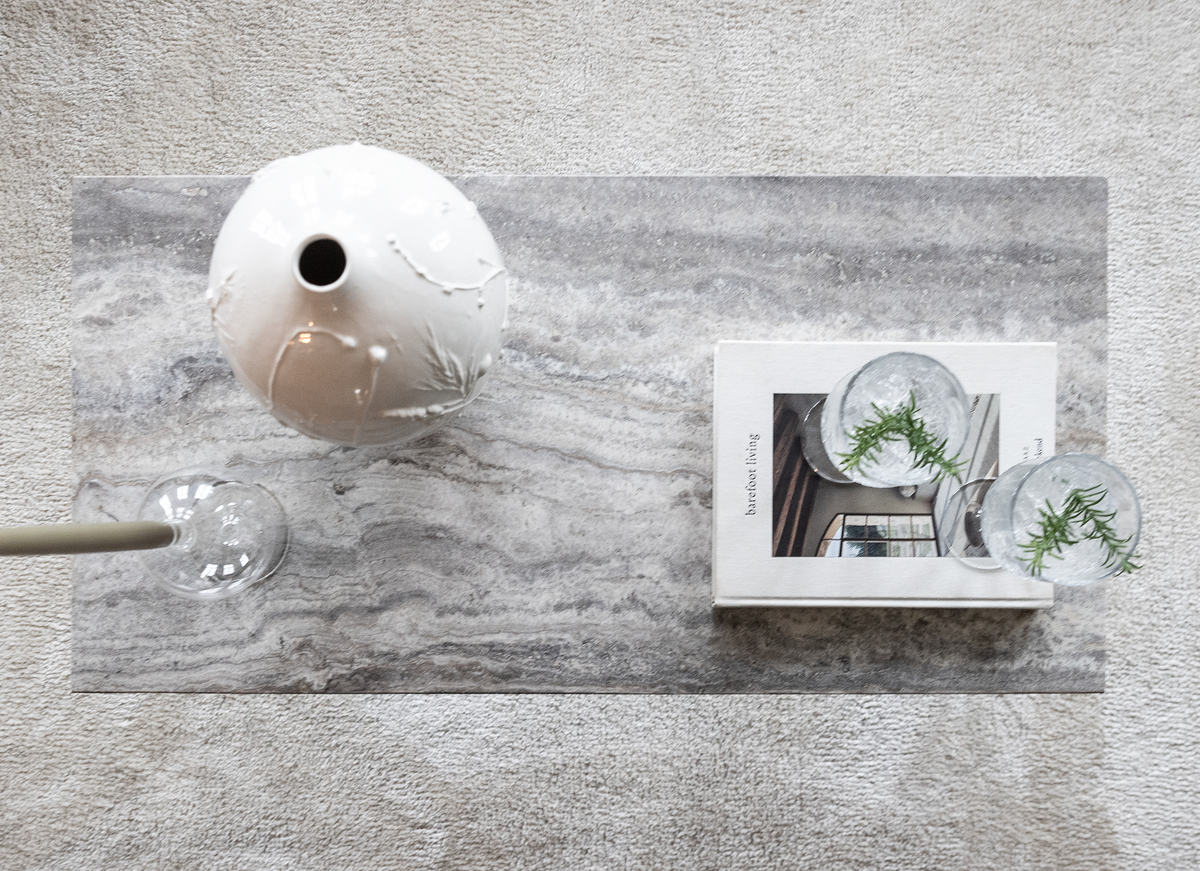 Top view af et gråt stenbord med en hvid keramisk vase, bogen ”barefoot living” og to klare glas med urtekviste på et lyst tæppe.