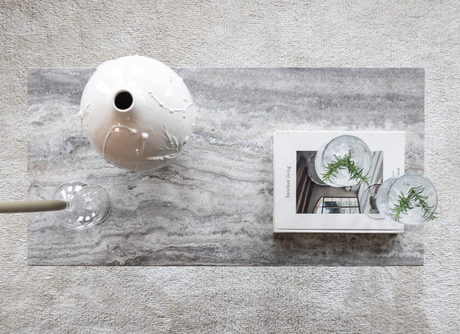 Top view af et gråt stenbord med en hvid keramisk vase, bogen ”barefoot living” og to klare glas med urtekviste på et lyst tæppe.
