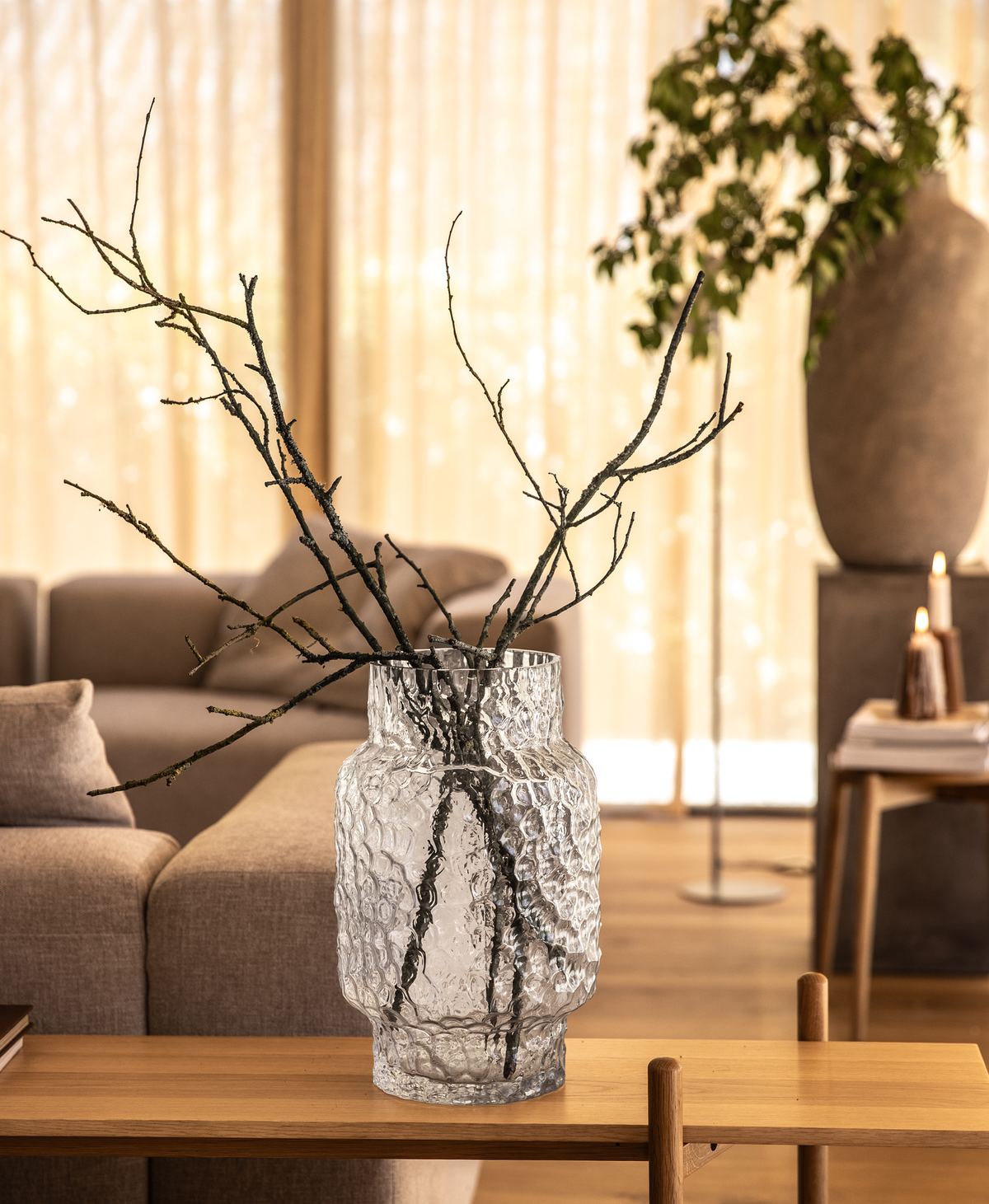 Struktureret klar glasvase med bare grene på et træbord i en varm, lys stue; beige sofa, gardiner og tændte lys i baggrunden.