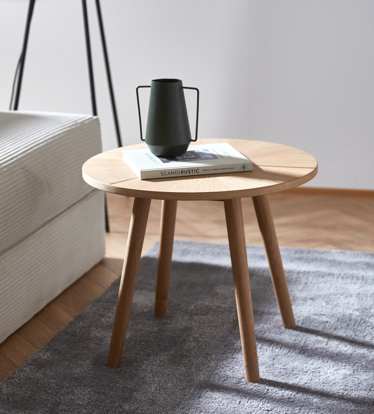 Rundt sidebord i eg på gråt tæppe ved siden af sofa, med mørk kande/vase oven på en bog med titlen SCANDIRUSTIC.