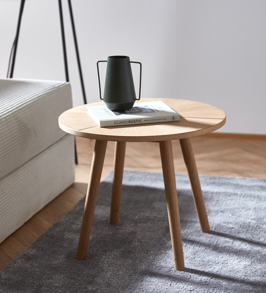 Rundt sidebord i eg på gråt tæppe ved siden af sofa, med mørk kande/vase oven på en bog med titlen SCANDIRUSTIC.