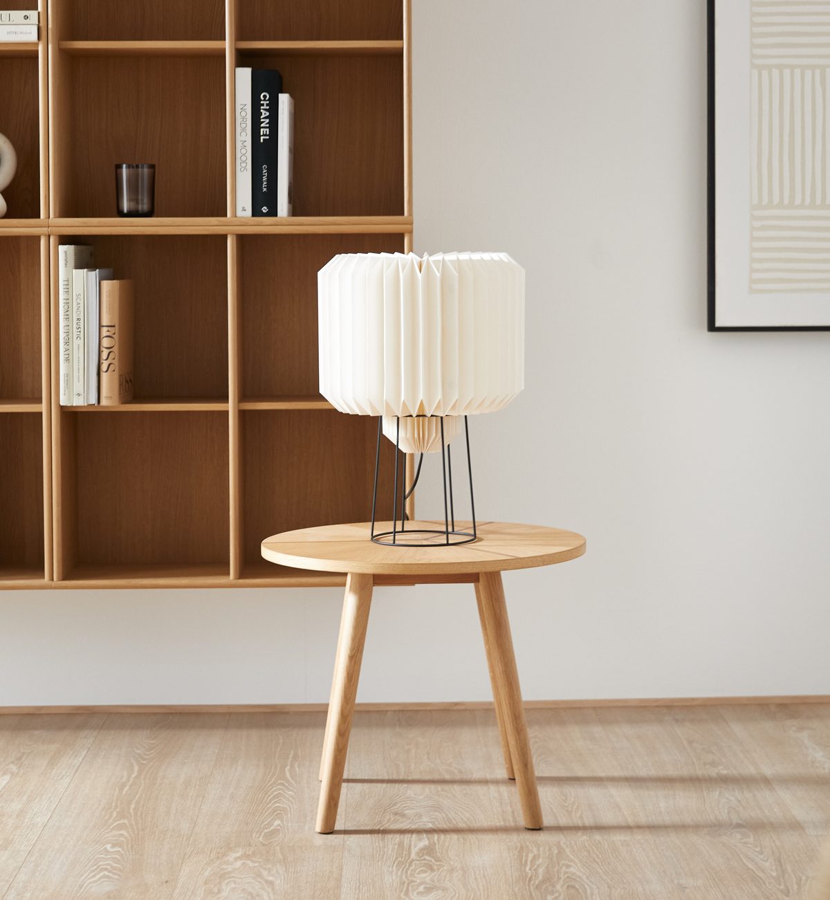 Lys stue med rundt sidebord i egetræ og hvid plisseret bordlampe; i baggrunden åben reol med bøger, bl.a. Chanel, og minimalistisk kunst.