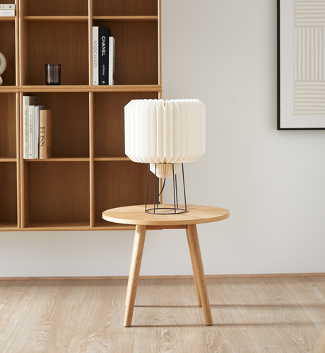 Lys stue med rundt sidebord i egetræ og hvid plisseret bordlampe; i baggrunden åben reol med bøger, bl.a. Chanel, og minimalistisk kunst.
