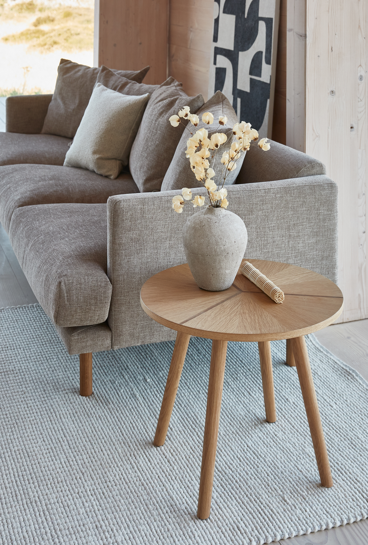 Lyst, skandinavisk stuehjørne med grå stofsofa med puder, et rundt sidebord i eg med en sten-vase med tørrede blomster, på et lyseblåt tekstureret tæppe.