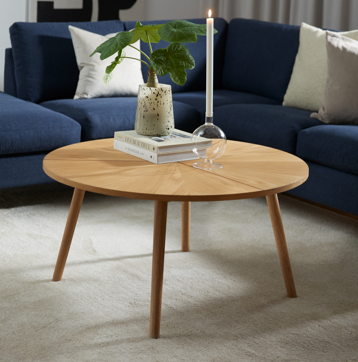 Rundt sofabord i eg med bøger—en med titlen ”LIVE BEAUTIFUL”—en spættet vase med grønne blade og et tændt stagelys; blå sofa og lyst tæppe i en lys stue.