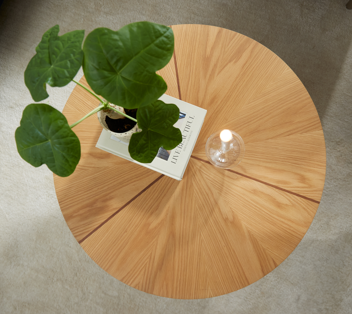 Rundt sofabord i lys egetræsfiner med radiært mønster, en grøn potteplante, en glaslysestage og bogen ”LIVE BEAUTIFUL” ovenpå.