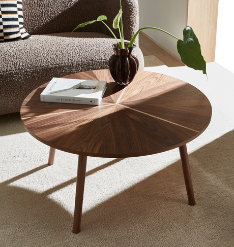 Rundt valnøds-sofabord med solstrålemønster, mørk vase med grøn plante og bogen The Kinfolk Home på en lys tæppe ved siden af en tekstureret sofa.