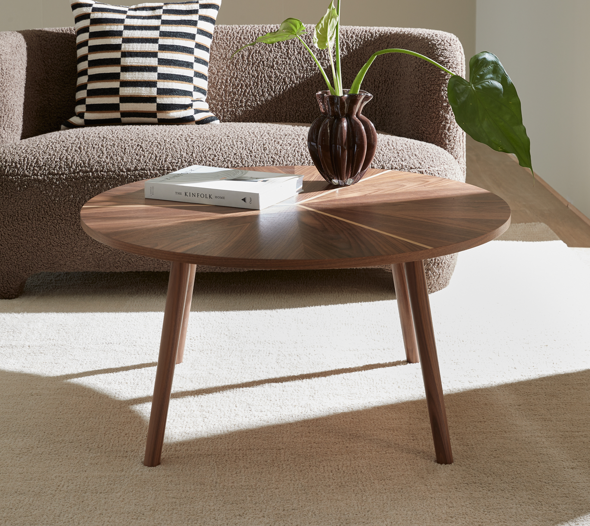 Rundt valnød sofabord med “The Kinfolk Home”, mørk glasvase og grøn plante på beige tæppe foran brun, tekstureret sofa.