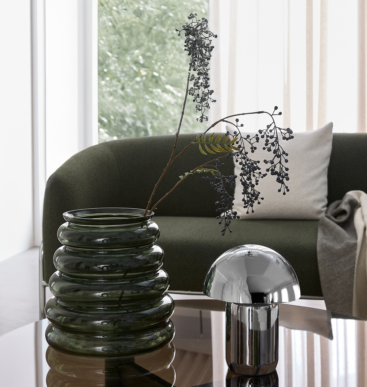 Nærbillede af en moderne stuescene med en riflet mørkegrøn glasvase med grene og en krom svampelampe på et blankt sofabord foran en grøn sofa med pude.