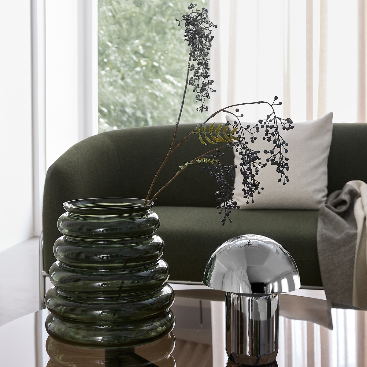 Grøn glasvase med gren og krom svamplampe på sofabord foran mørkegrøn sofa med pude og plaid i en lys stue.