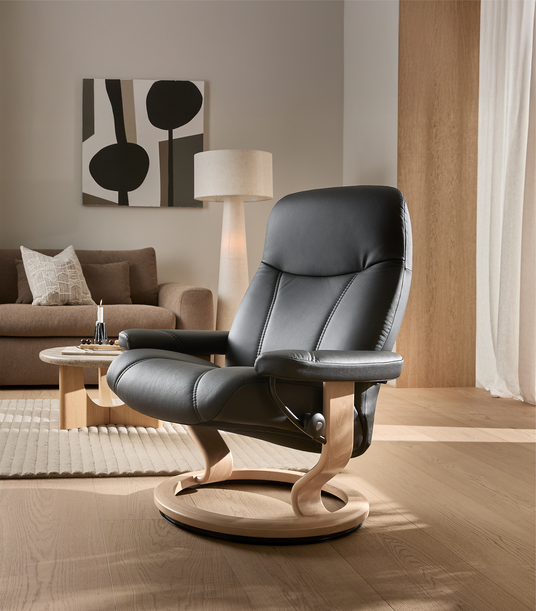 Sort læderlænestol med lys træsokkel i en lys stue med beige sofa, gulvlampe og abstrakt vægkunst.
