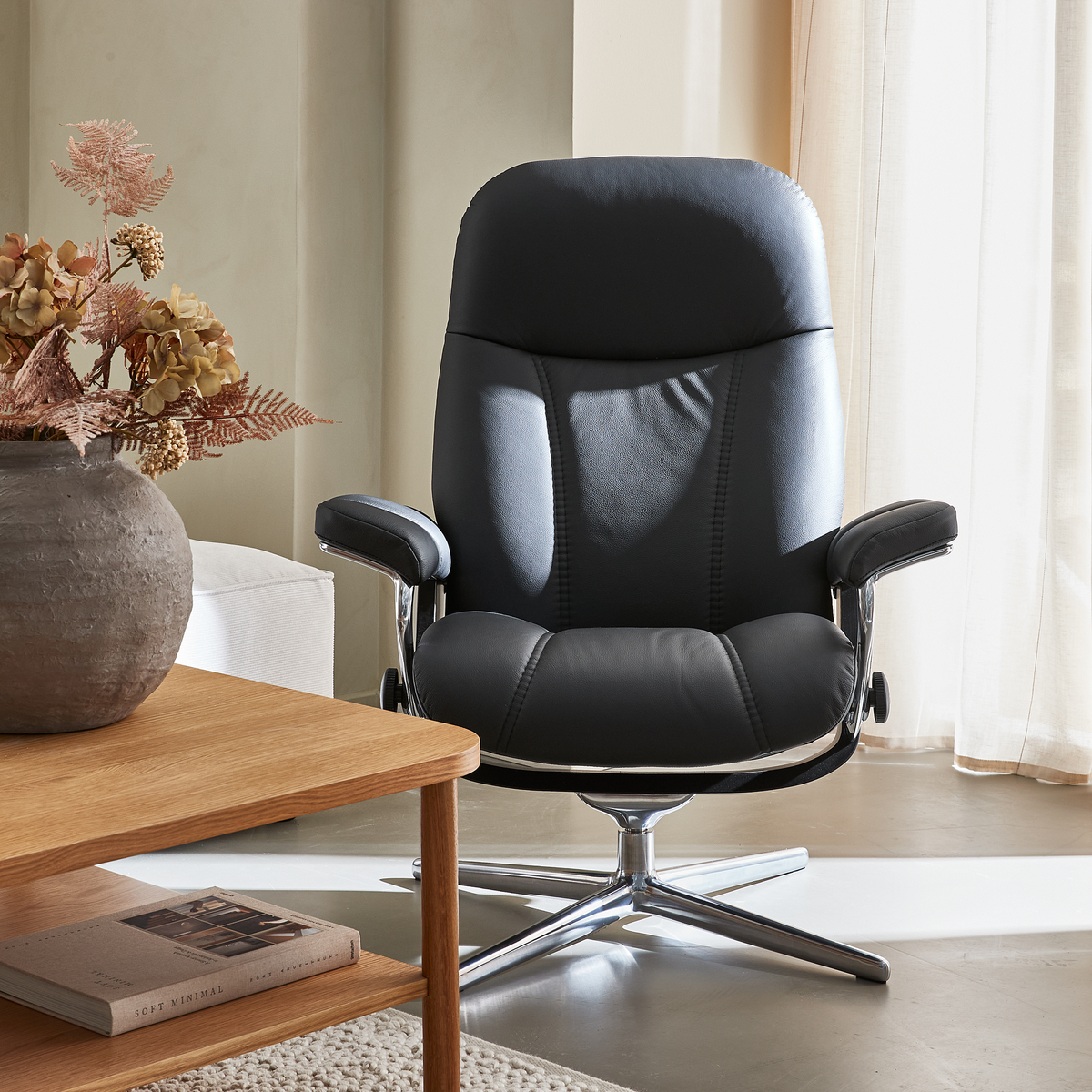Sort læderlænestol med krom drejefod ved siden af et egetræsbord med vase med tørrede blomster og bogen Soft Minimal i en lys stue.
