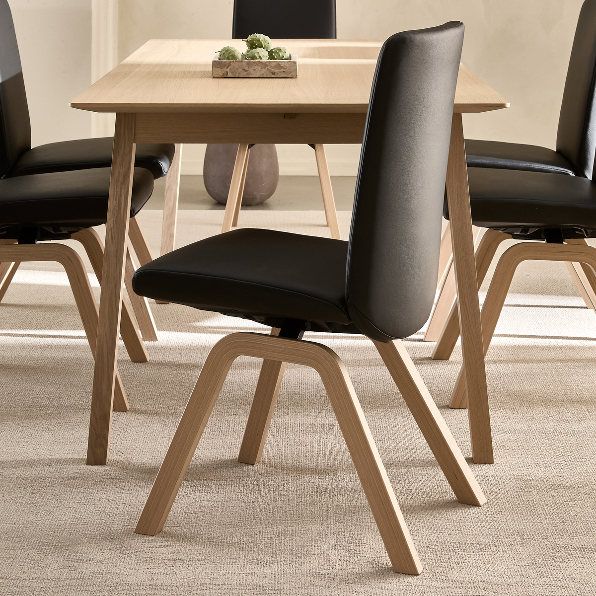 Spisestol i sort læderlook med bøjetræben ved et lyst træbord på beige tæppe, med en lille bakke med grønne sukkulenter.
