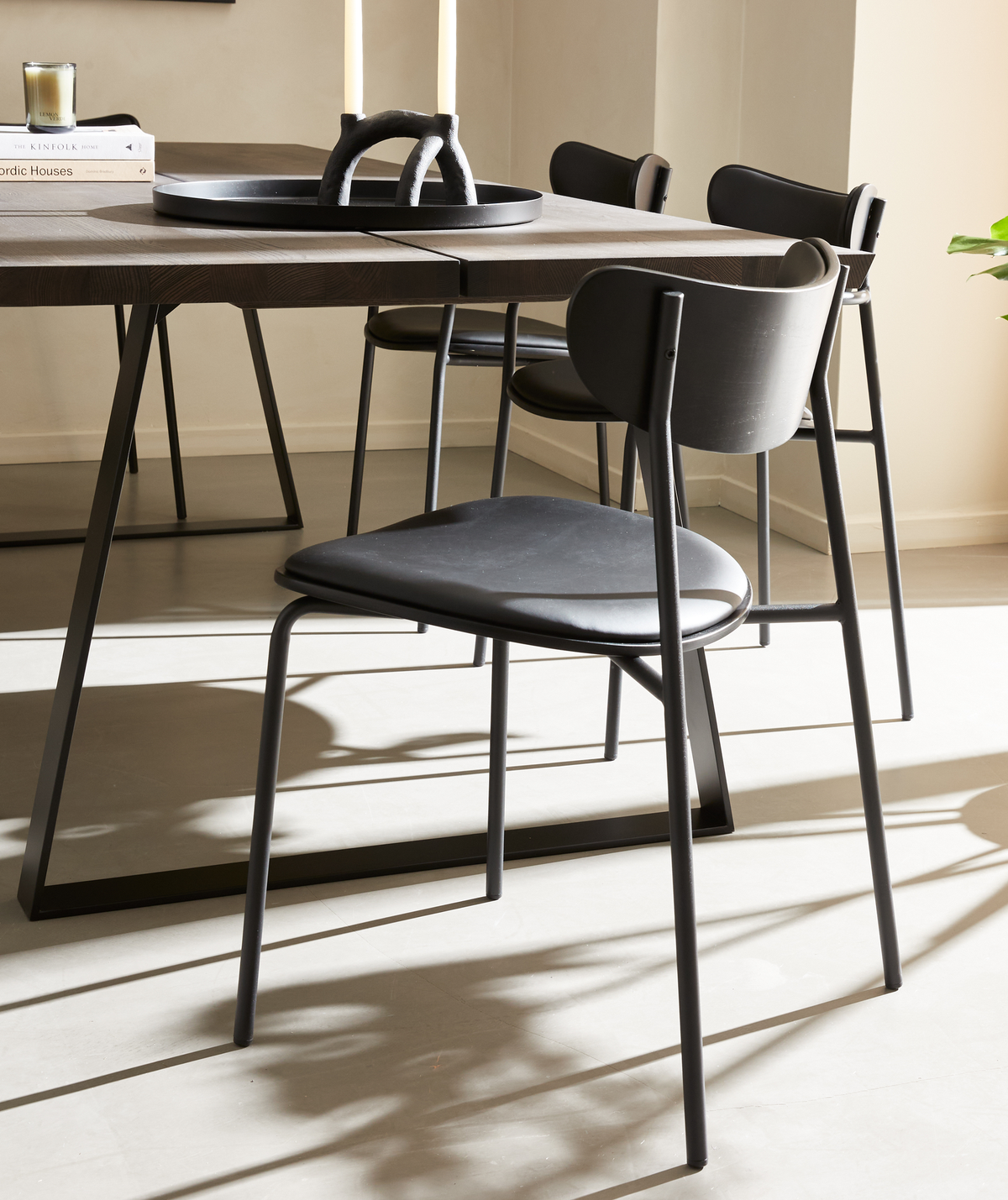 Sort spisebordsstol med buet ryg og slanke metalben ved et mørkt træbord med metalstel; på bordet står et fad med to lys i en skulpturel lysestage samt bøgerne Nordic Houses og Kinfolk i blødt dagslys.