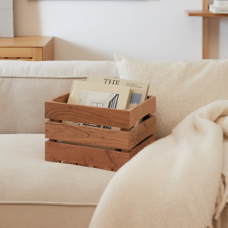 En trækasse med magasiner, herunder et med titlen “THE” og underrubrikken “New homes in residential buildings”, står på en beige sofa med pude og lys plaid.