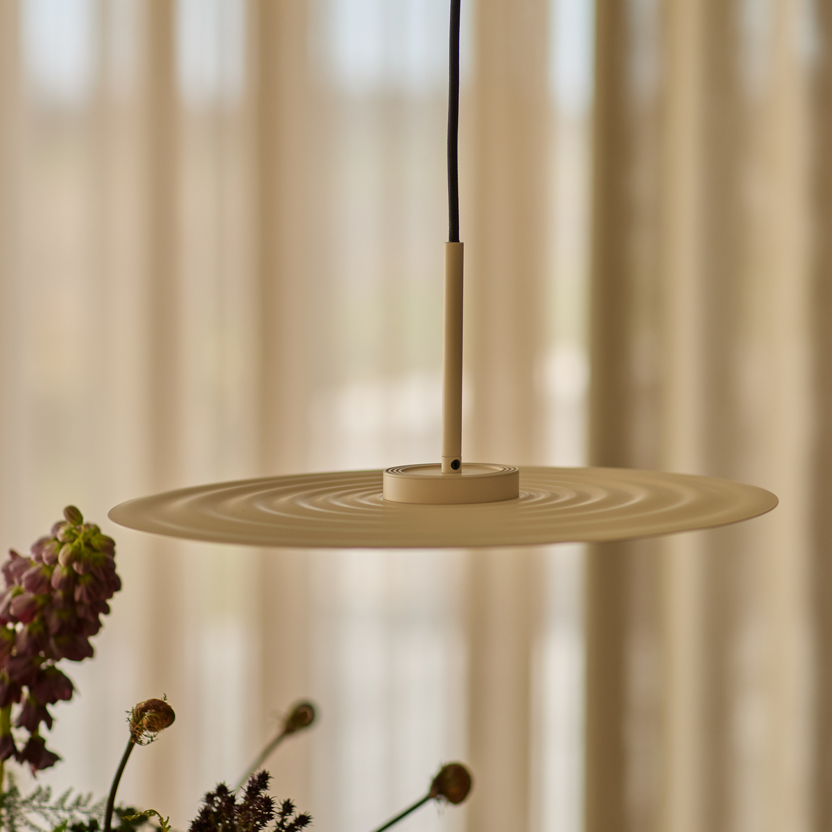 Beige, skiveformet pendellampe med koncentriske riller hænger over blomster i et blødt oplyst rum med gardiner.