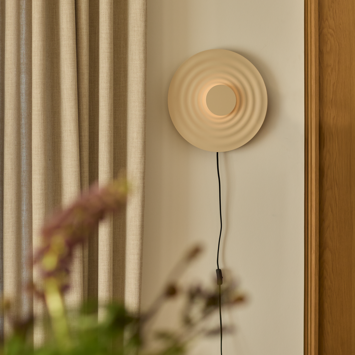 Væglampe med rund bølget skærm i beige, tændt, ved siden af beige gardin og træpanel; sort ledning hænger ned.