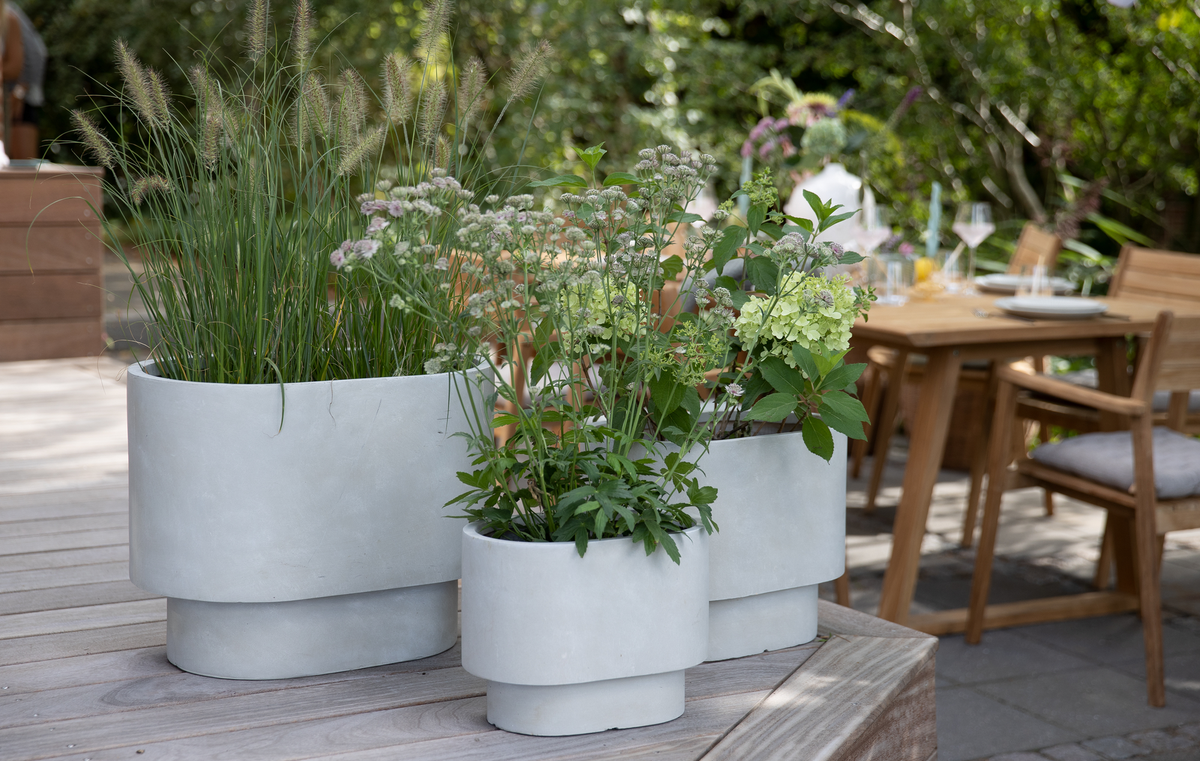 Tre lysegrå moderne plantekrukker med grønne planter på en træterrasse, med dækket havebord i baggrunden.