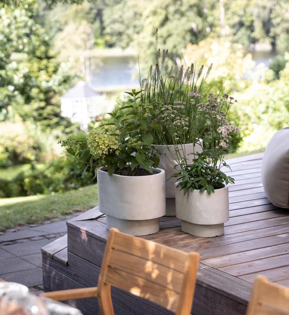 Tre lysegrå plantekrukker med grønne planter på en træterrasse med sløret have og sø i baggrunden.
