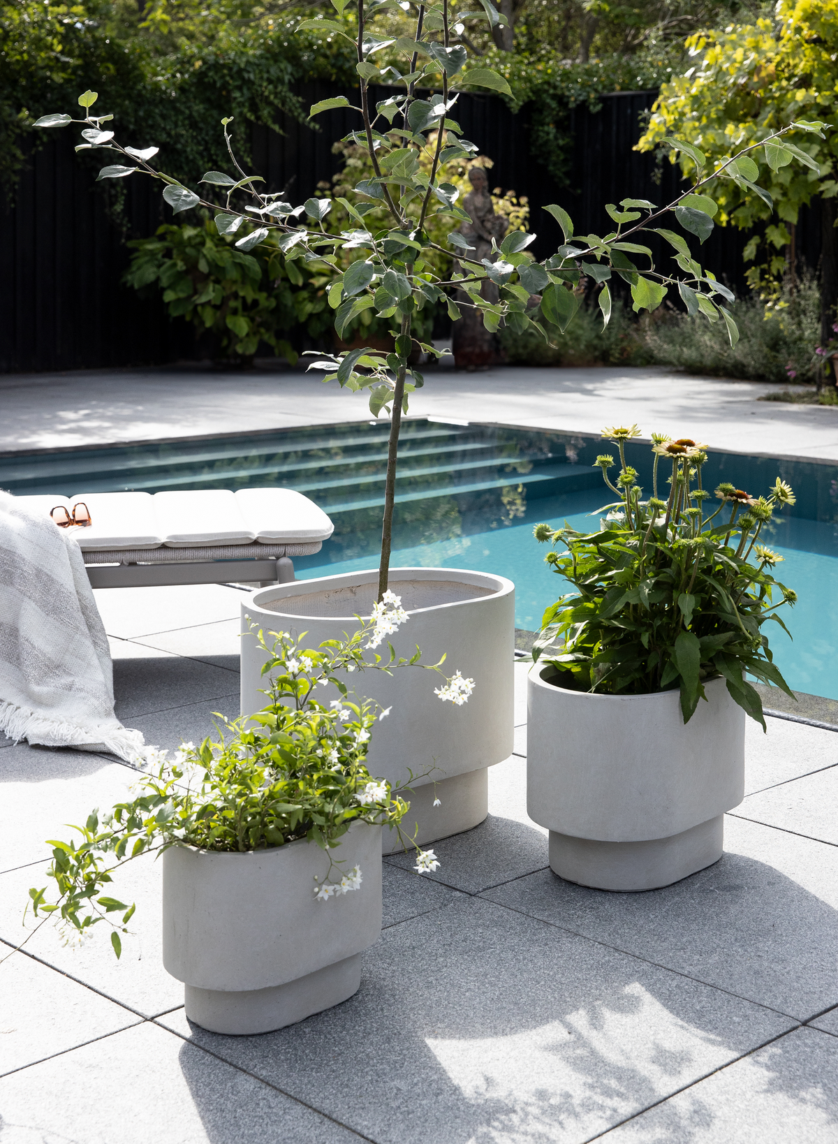 Tre lysegrå moderne plantekrukker med grønne planter ved en pool på en solrig flisebelagt terrasse; liggestol med plaid i baggrunden.