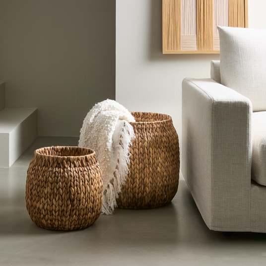 To flettede kurve i naturmaterial ved siden af en lys beige sofa, hvor den større kurv rummer et hvidt, blødt plaid.