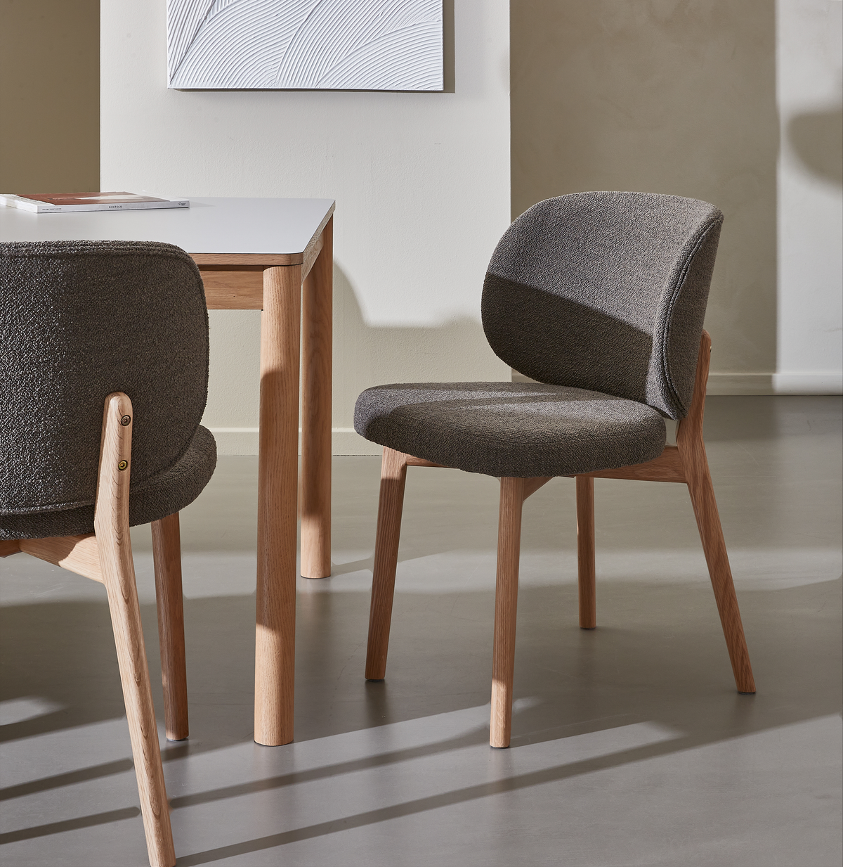 To grå polstrede stole med lyse træben ved et bord i et minimalistisk, lyst interiør med vægkunst og solskygger.