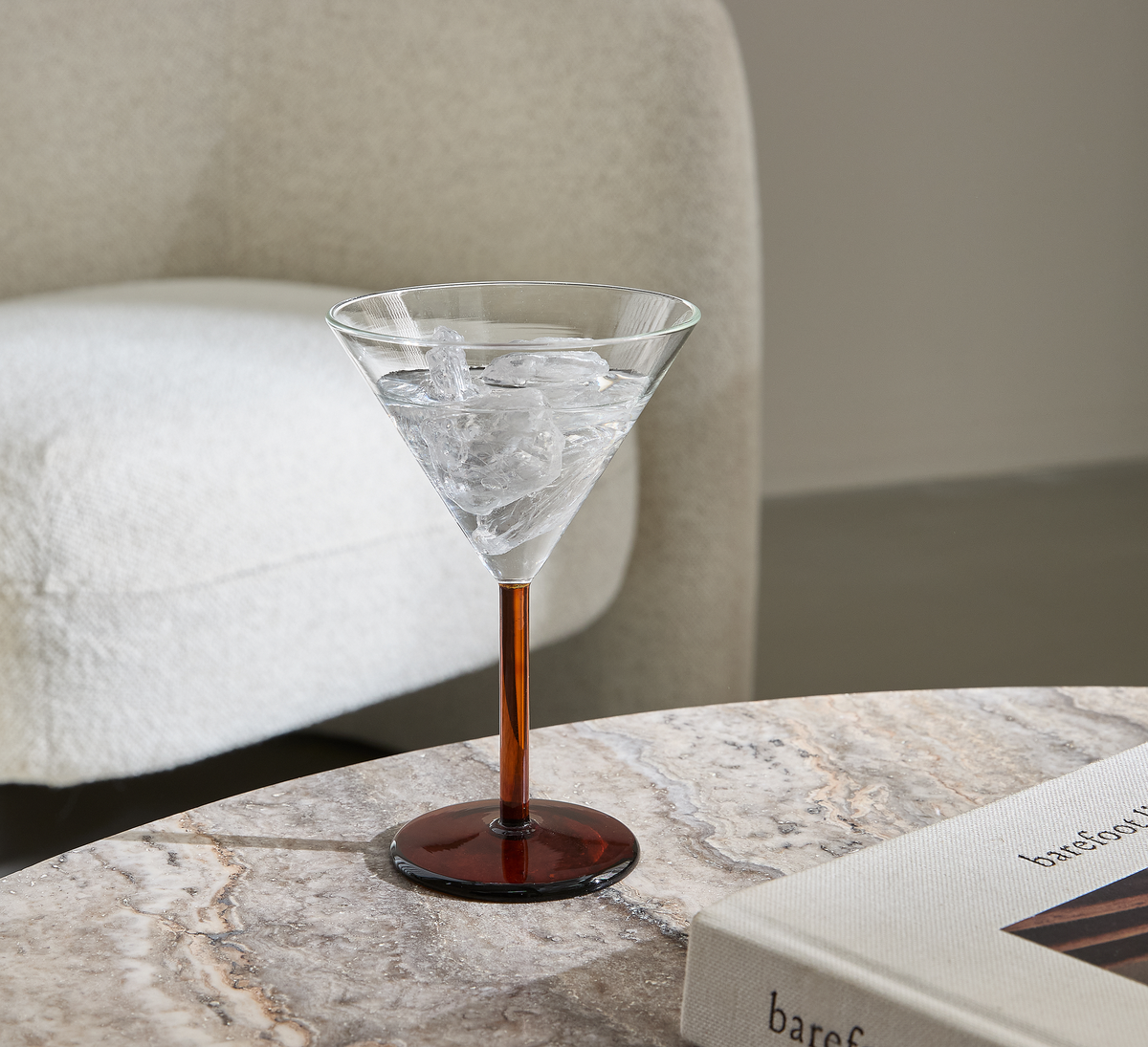 Martiniglas med is på et marmorbord ved siden af en beige lænestol og en coffeetable-bog med titlen ”baref…”.