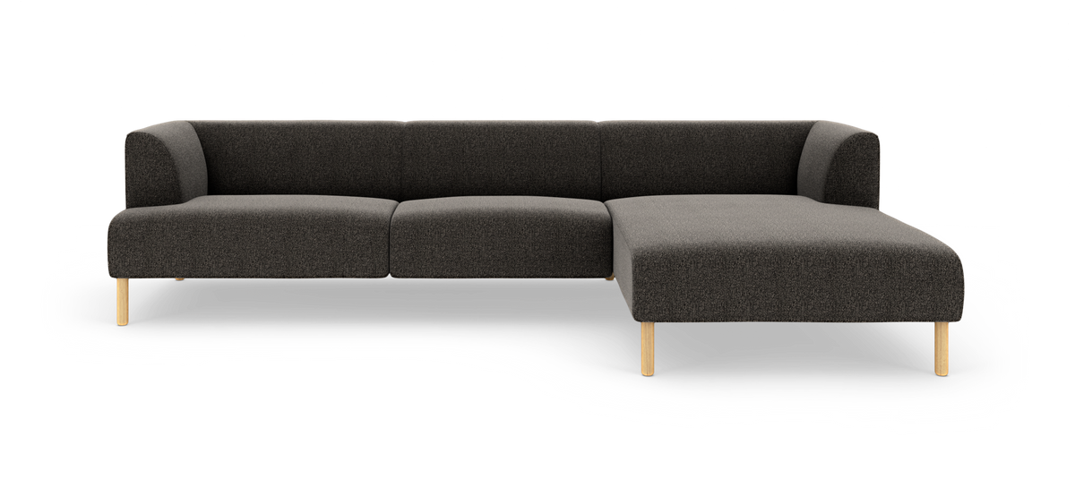 Mørkegrå L‑sofa med chaiselong til højre, afrundede hjørner og lyse træben, frilagt på hvid baggrund.