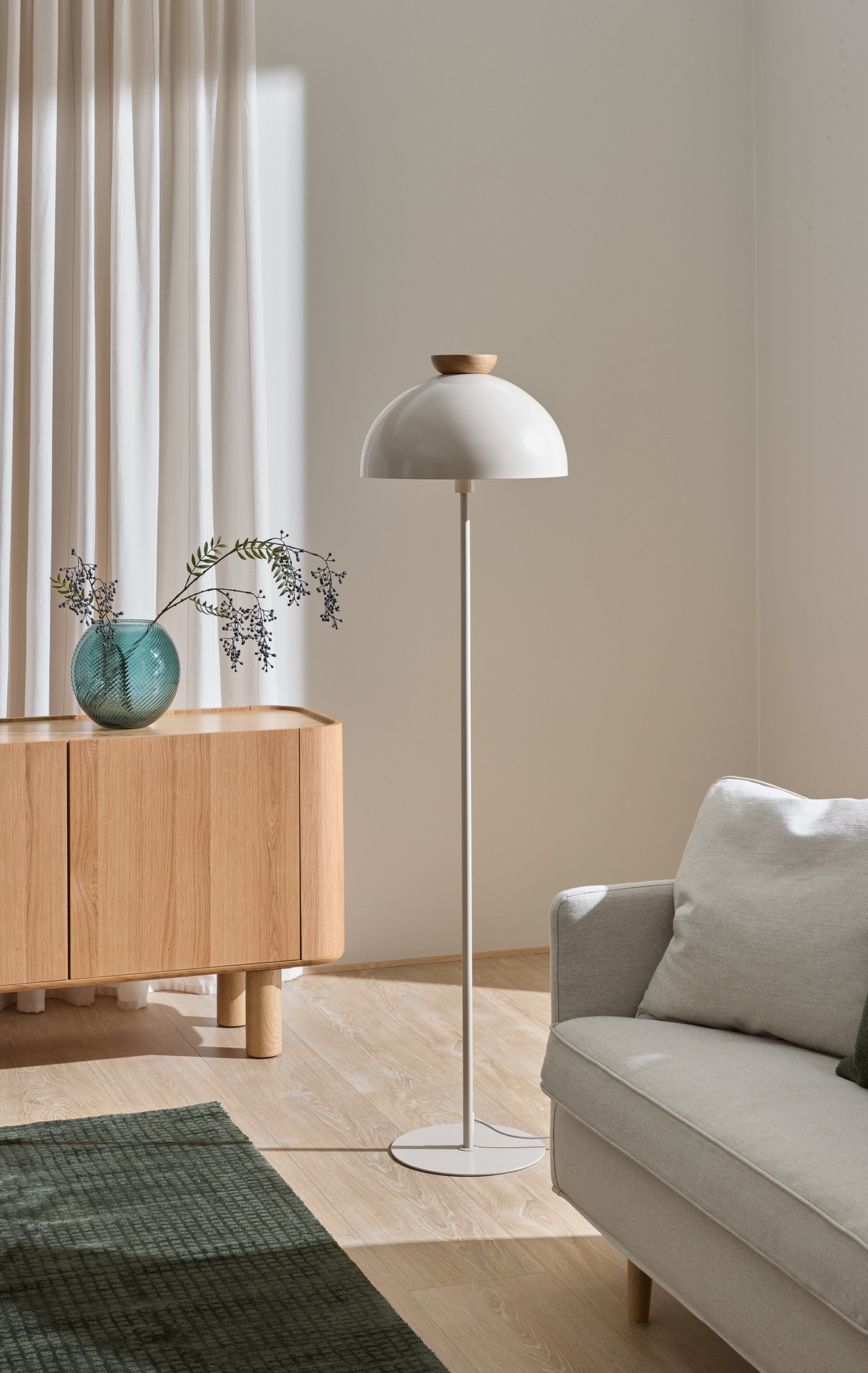 Lyst og minimalistisk hjørne af stue med hvid gulvlampe, egetræsskænk med turkis vase og grene, lysegrå sofa og grøn måtte i blødt dagslys fra vindue med gardiner.