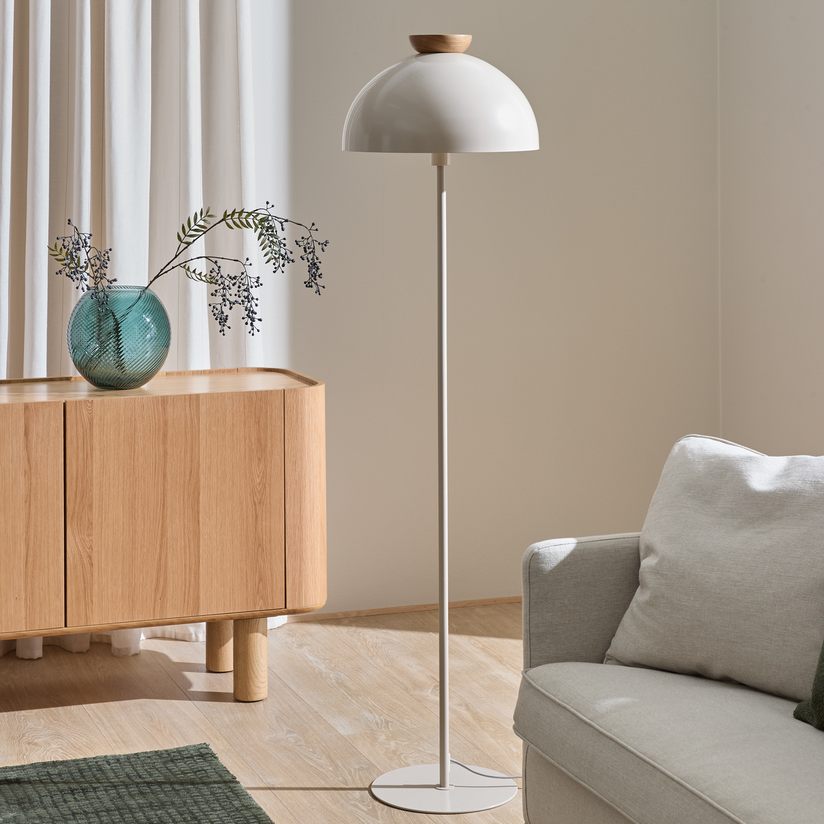 Minimalistisk stuekrog med hvid gulvlampe, skænk i egetræ med grøn glasvase og grene, lysegrå sofa og grønt tæppe i blødt dagslys.