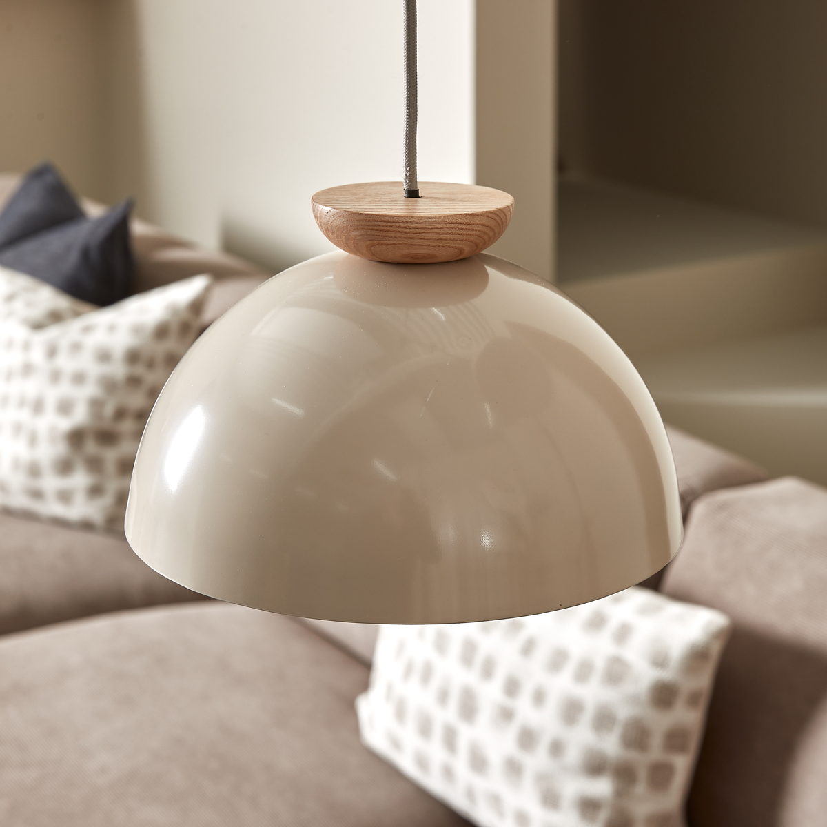 Beige kuppelformet pendellampe med trædetalje over en neutral sofa med mønstrede puder.