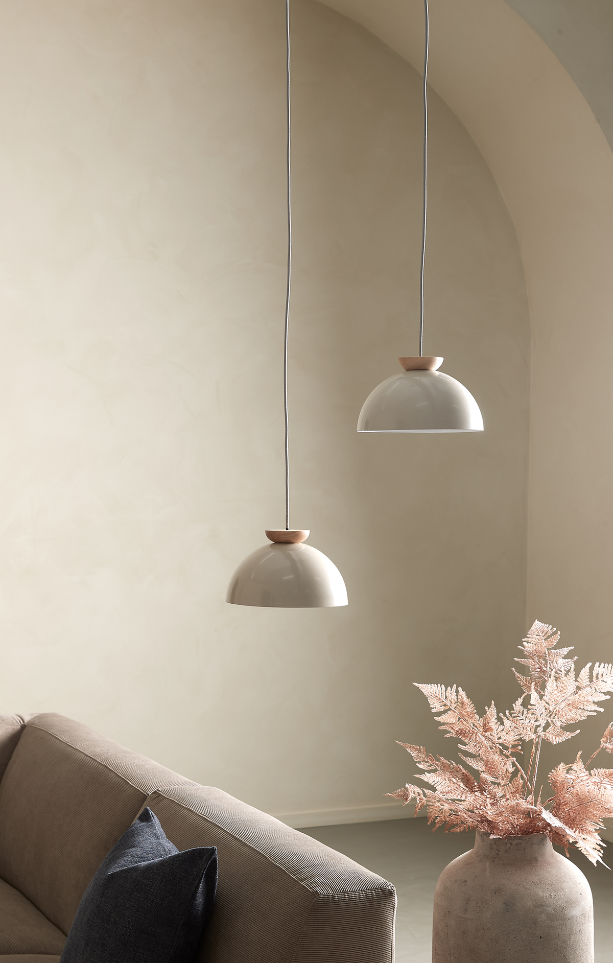 Lyst og minimalistisk stue med to beige kuppelformede pendellamper, en beige sofa med mørk pude og en stor stenvase med tørrede lyserøde grene foran en buet væg.