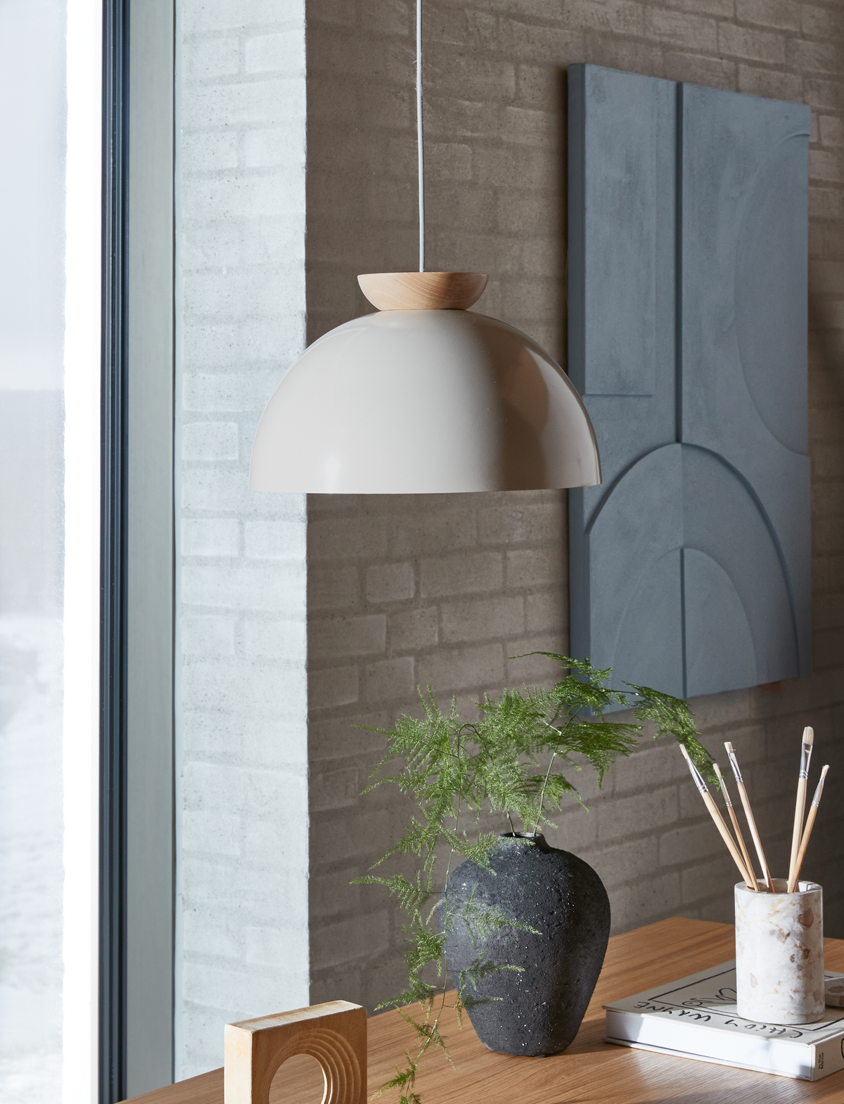 Beige pendellampe med trædetalje over et træbord med sort vase og grønne kviste ved et vindue.