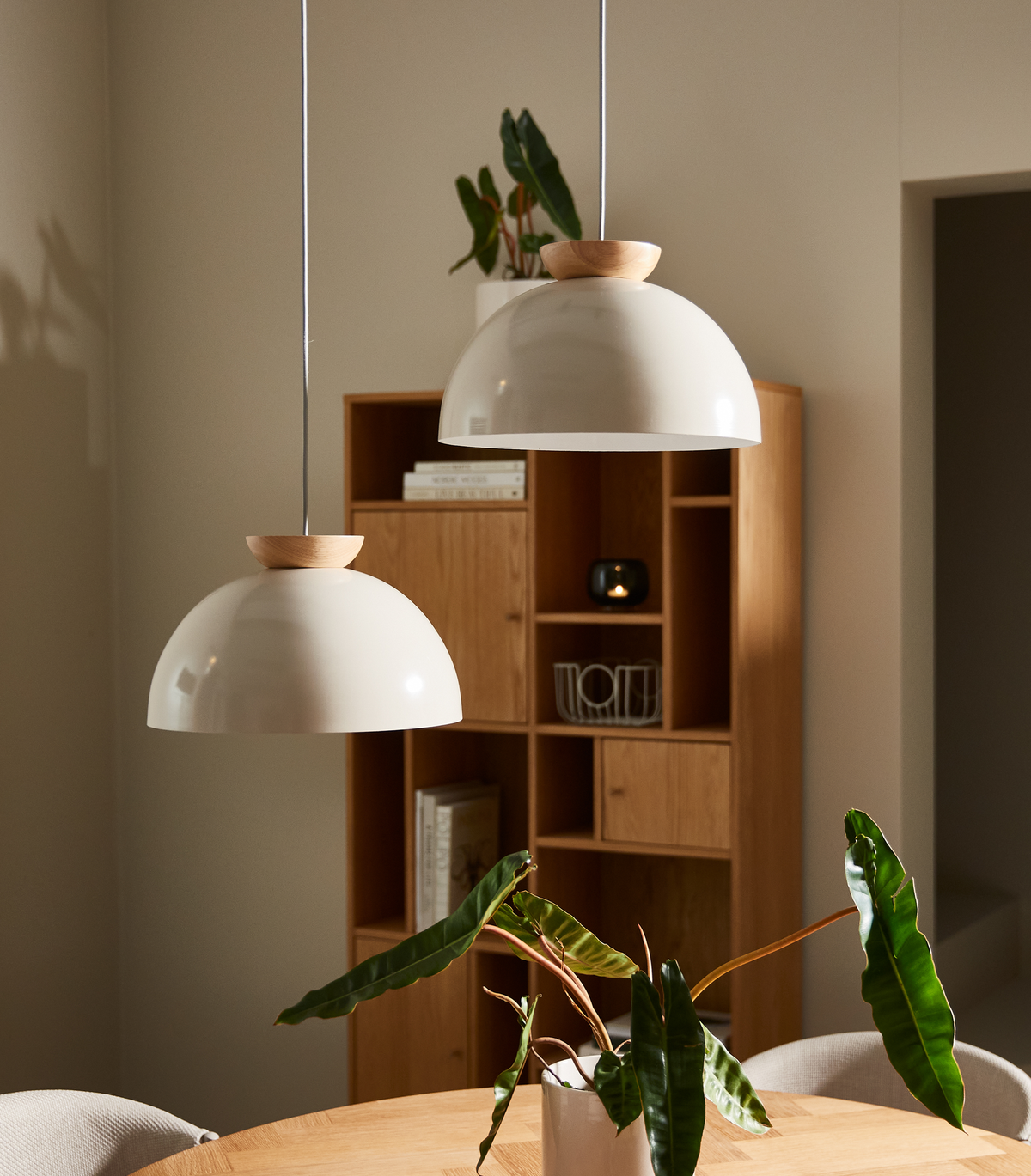 To hvide pendler med trædetaljer over et rundt spisebord, grøn plante i vase og egetræshylde i baggrunden.