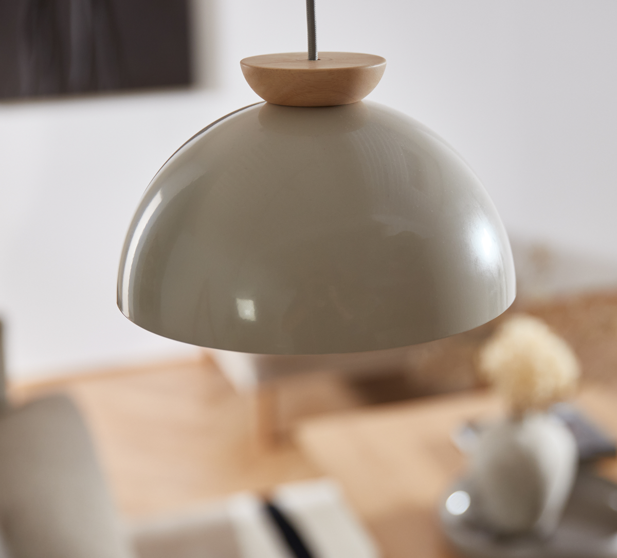 Beige kuppelformet pendel med trædetalje hænger over et lyst spisemiljø i naturlige nuancer.