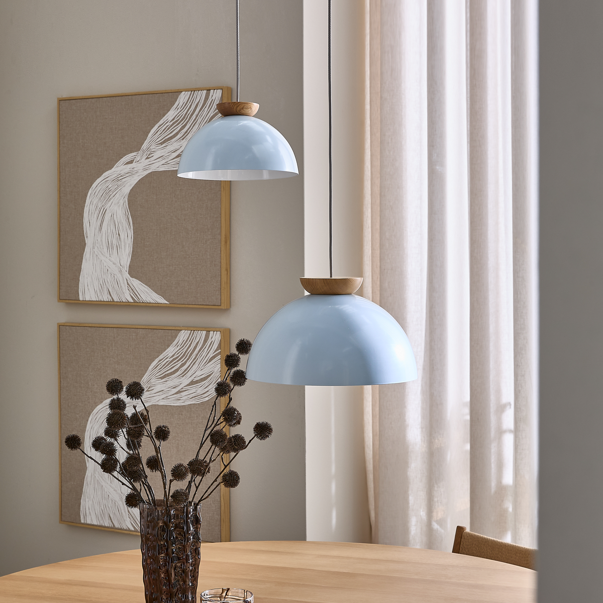 To lyseblå kuppelformede pendler med trædetaljer over et lyst træbord, vase med tørrede grene, beige gardiner og abstrakt vægkunst i rolig skandinavisk indretning.