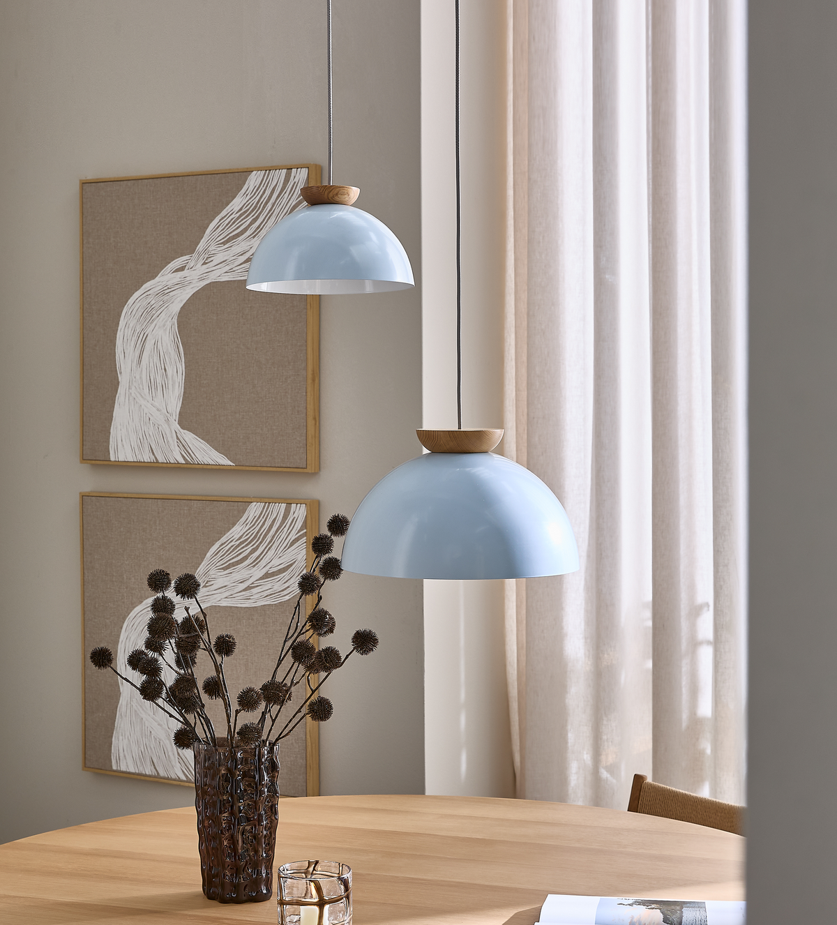 To lyseblå pendler med trædetaljer over et rundt bord i lyst træ, vase med tørrede grene, beige gardiner og abstrakt vægkunst i neutrale toner.
