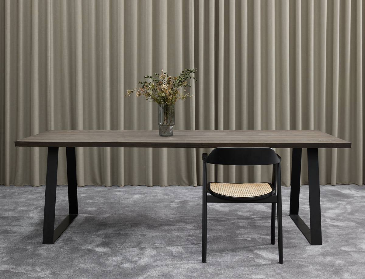 Minimalistisk spisescene med mørkt træbord på sorte bukke, en sort stol med rattan-sæde og en vase med tørrede blomster foran grå gardiner og tæppe.