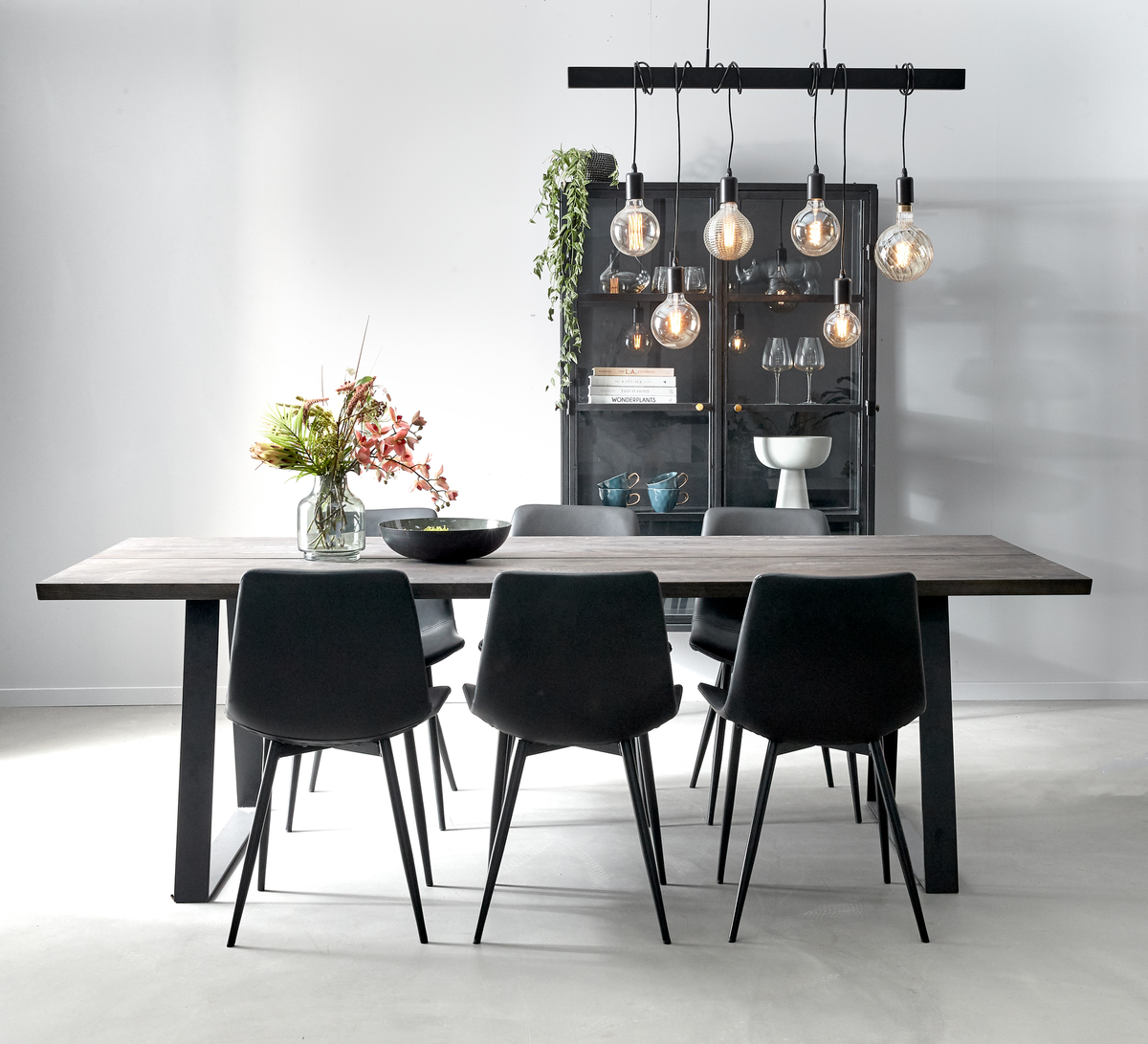 Moderne spiseplads med sort pendellampe med synlige pærer over et rektangulært træbord, seks mørke stole og et sort vitrineskab i baggrunden; vase med blomster og grøn plante som detaljer.