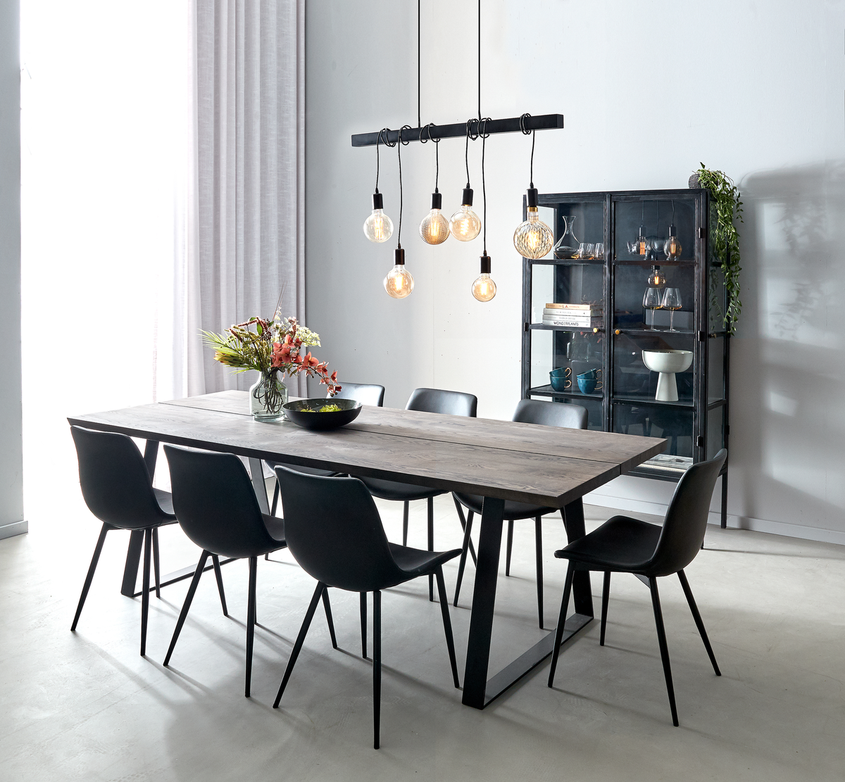 Spisestue med lang rektangulær træbordplade, otte sorte stole, sorte pendellamper med varme pærer og et metalskab med glas; vase med blomster på bordet.