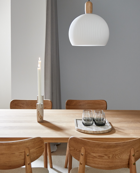 Lyst spisestue med træbord og fire træstole, hvid pendellampe, to tændte lys og en bakke med glas på bordet.