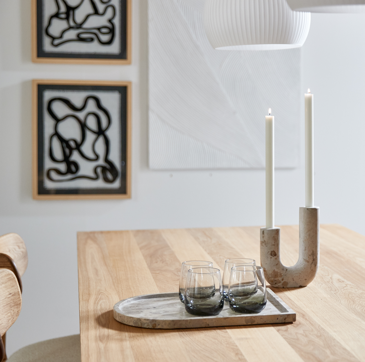Lyst spisebord i træ med marmorbakke med fire glas og U‑formet lysestage med to tændte stearinlys; abstrakt vægkunst og hvid pendel i baggrunden.