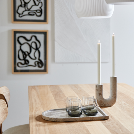 Lyst spisebord i træ med marmorbakke med fire glas og U‑formet lysestage med to tændte stearinlys; abstrakt vægkunst og hvid pendel i baggrunden.